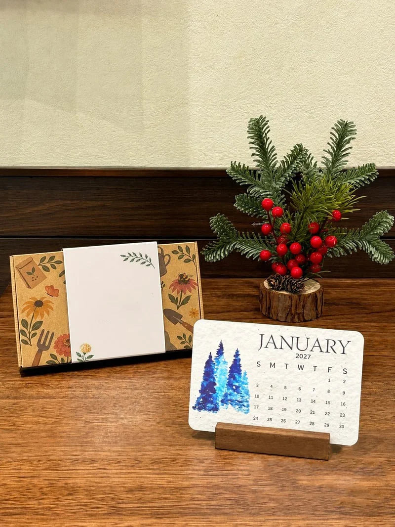 January 2027 calendar page displayed on a wooden stand beside botanical gift packaging